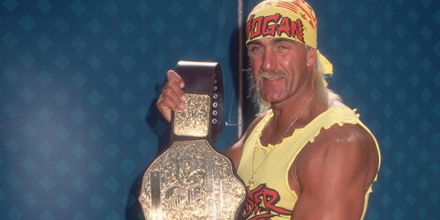 Hulk Hogan showing off his championship belt.  Hogan wears a costume consisting of a yellow 'HOGAN' bandana, a yellow top, a weightlifter belt, red spandex pants, and yellow cowboy boots. 