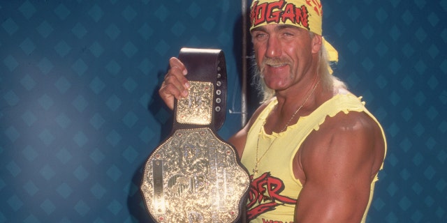 Hulk Hogan showing off his championship belt.  Hogan wears a costume consisting of a yellow 'HOGAN' bandana, a yellow top, a weightlifter belt, red spandex pants, and yellow cowboy boots. 