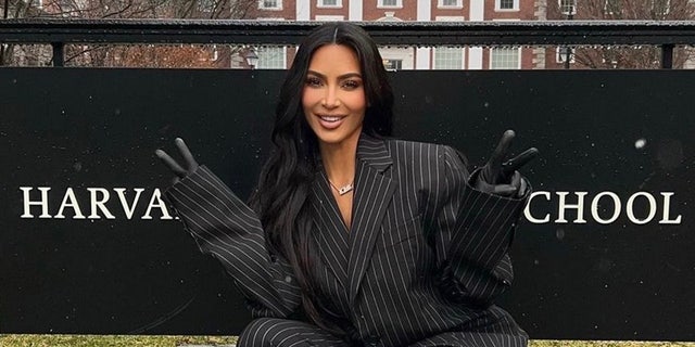 Kim Kardashian in a black and white pinstripe suit gives to peace signs in front of a sign that says 'Harvard Business School'