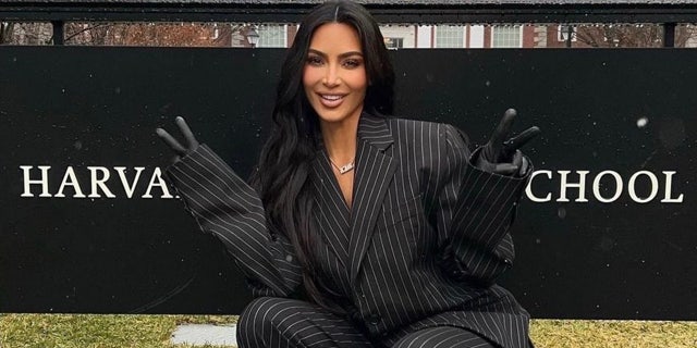 Kim Kardashian in a black and white pinstripe suit gives to peace signs in front of a sign that says 'Harvard Business School'