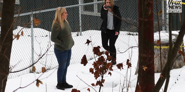 Two women stand in the snow at the back of a house while one of them uses a video camera.