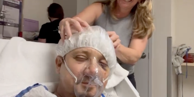 Renner is seen massaging his head while wearing a hair net, breathing medical mask, and lying on a hospital bed. 