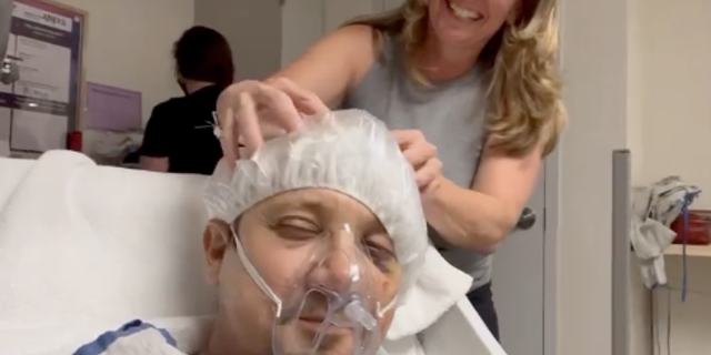 Renner é visto massageando a cabeça enquanto usava uma rede para o cabelo, respirando máscara médica e deitado em uma cama de hospital. 