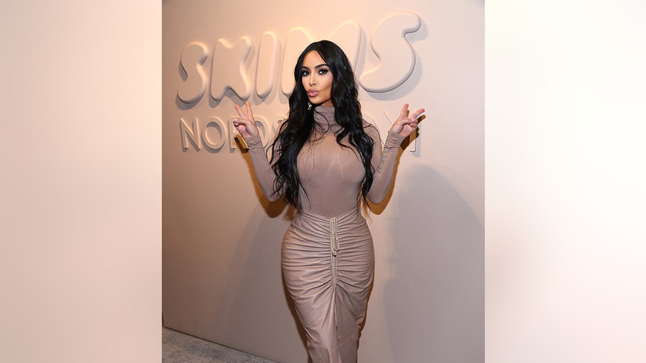 Kim Kardashian in a champagne top and skirt gives a kissy face and two peace signs in front of a SKIMS sign at Nordstrom