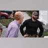 A man wearing a plaid shirt exits a police vehicle surrounded by officers.