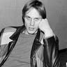 Tom Verlaine with his a fist to his face in a patterned leather jacket stares at the camera