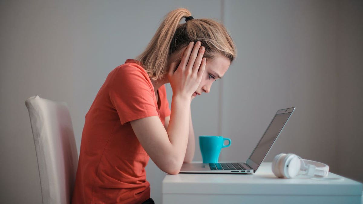 Woman in an orange shirt sitting at the table, staring at her laptop.