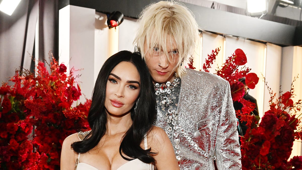 Megan Fox leans against Machine Gun Kelly in a glittery silver jacket and long chain, Fox wears a busty whit dress