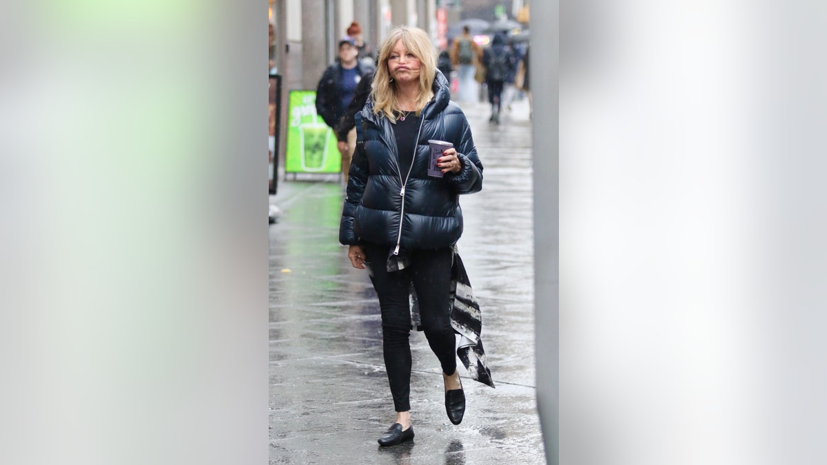 Goldie Hawn makes a funny duck-looking face gesture while holding a purple protein drink and sporting a stylish puffer jacket during a rainy morning in Manhattan’s Midtown area.