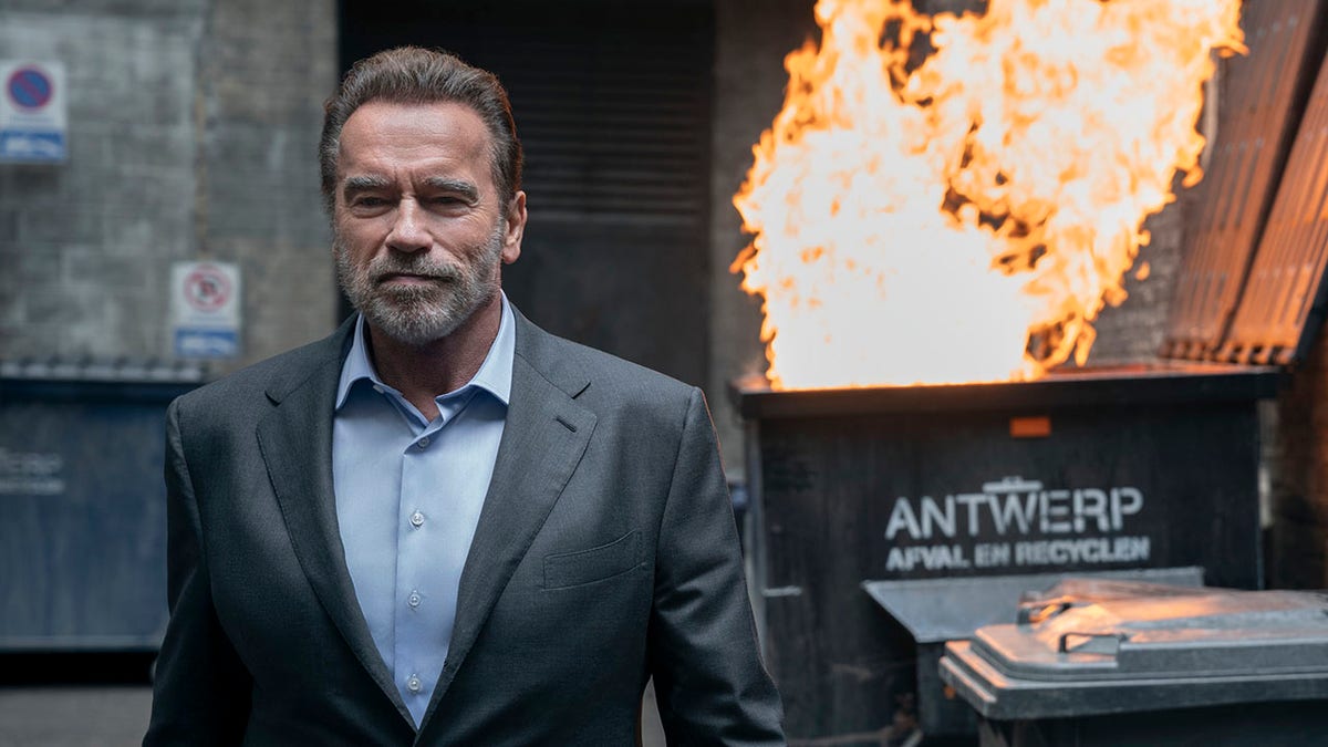 Arnold Schwarzenegger wears a blue button down and fitted grey suit while walking forward, behind him a dumpster fire is erupting in a photo from his new show "Fubar"