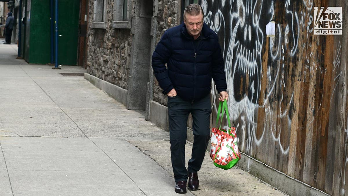 Alec Baldwin dressed in dark clothes has his hand in his pocket as he walks along the sidewalk