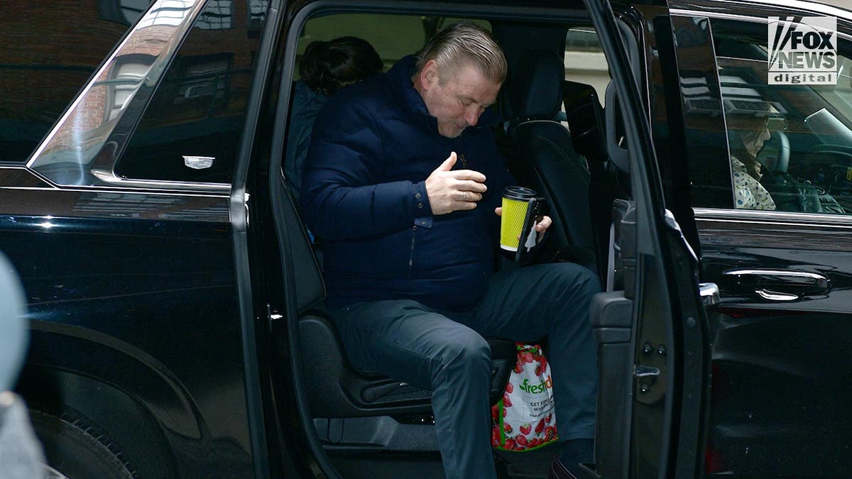 Alec Baldwin turns to get out of car with yellow cup of coffee in hand