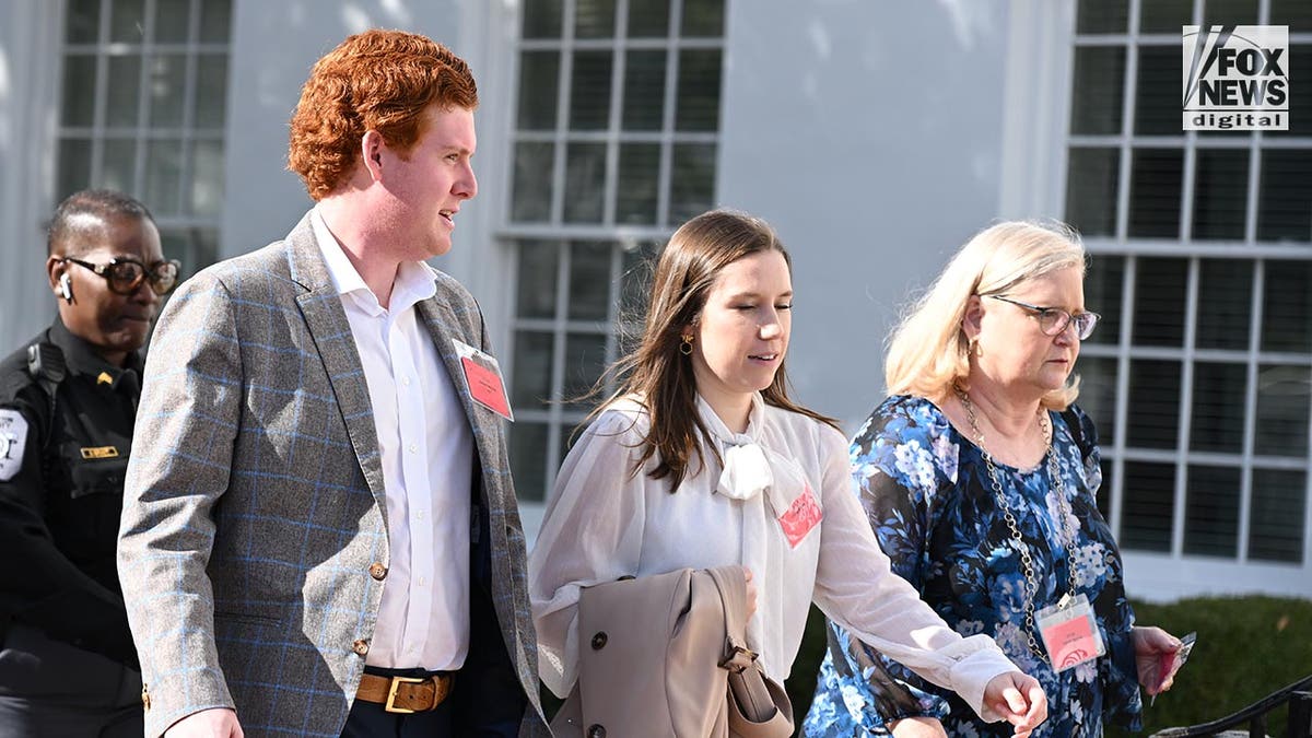 Buster in a grey blazer talks to Brooklyn, wearing a white blouse, Lynn walks alongside in a blue floral dress