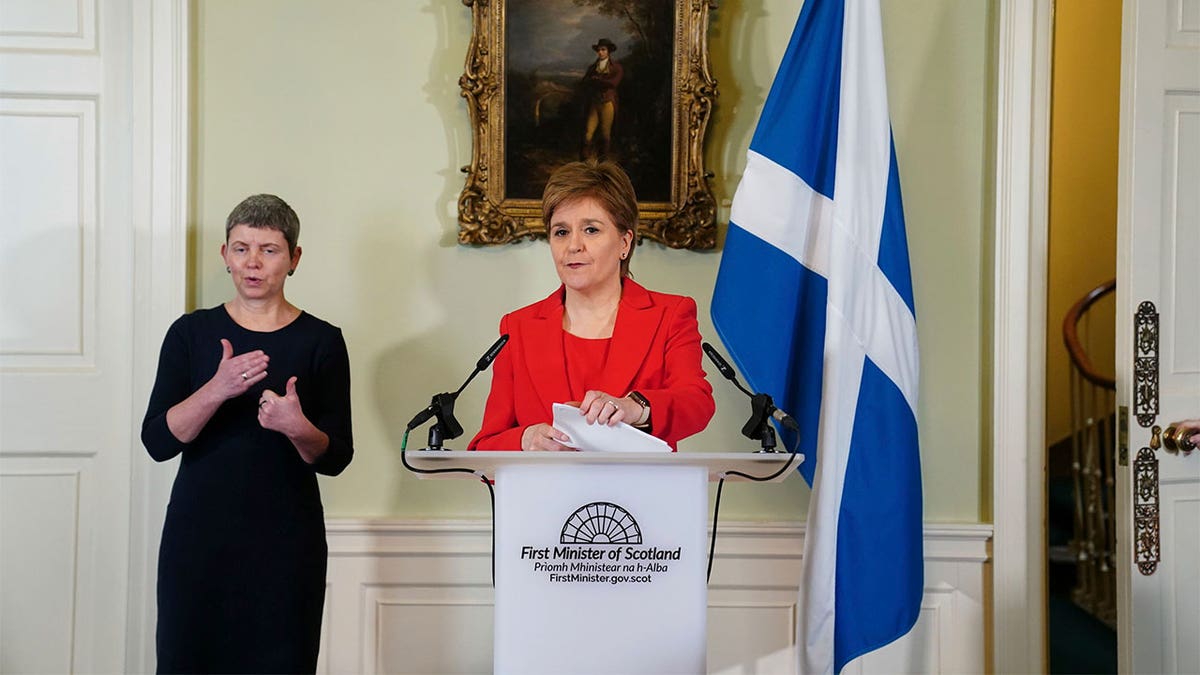 Nicola Sturgeon holding a press conference wearing a red blazer and standing in front of a white podium.