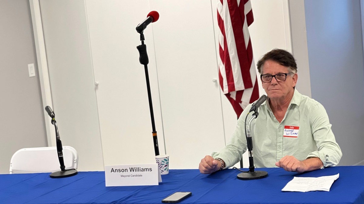 Anson Williams looking serious while sitting next to a blue table and an American flag