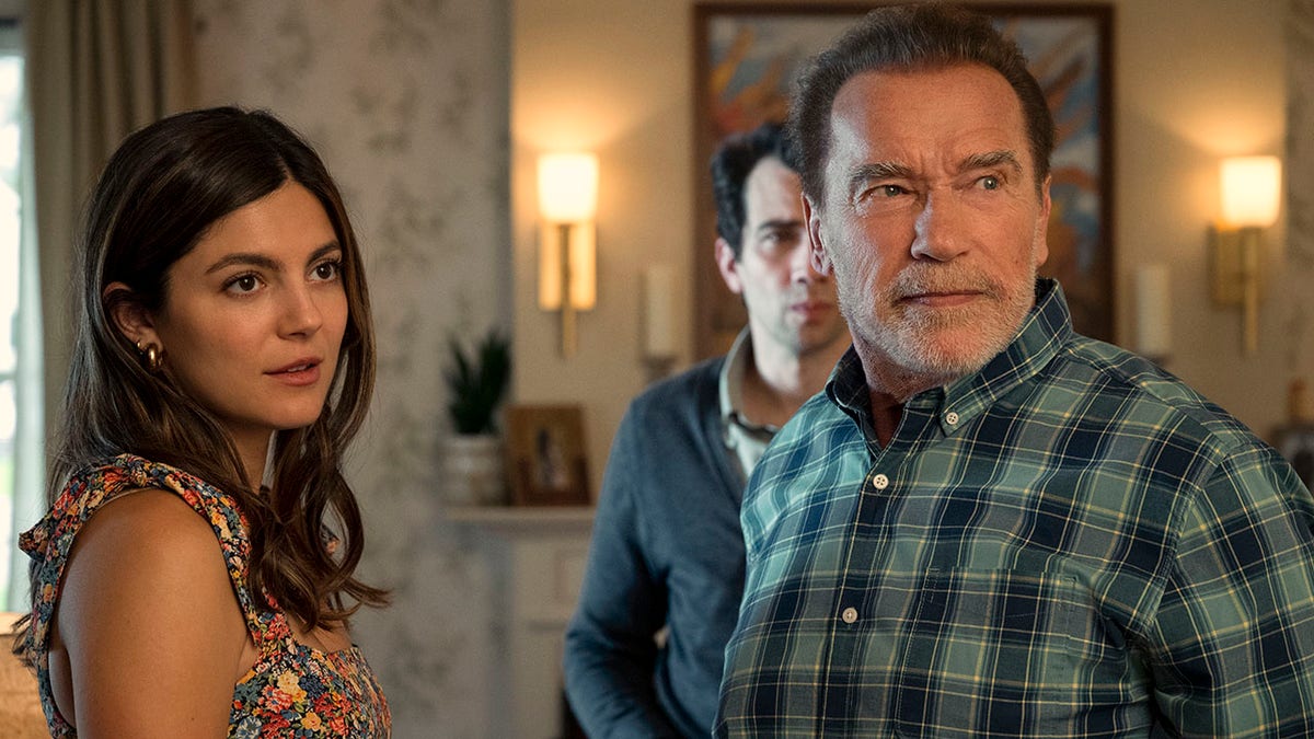 Monica Barbaro as Emma Brunner in a floral tank top looks off camera alongside Arnold Schwarzenegger as Luke Brunner in a plaid two-toned green shirt with actor Jay Baruchel as Carter Perlmutter in the background wearing a suit