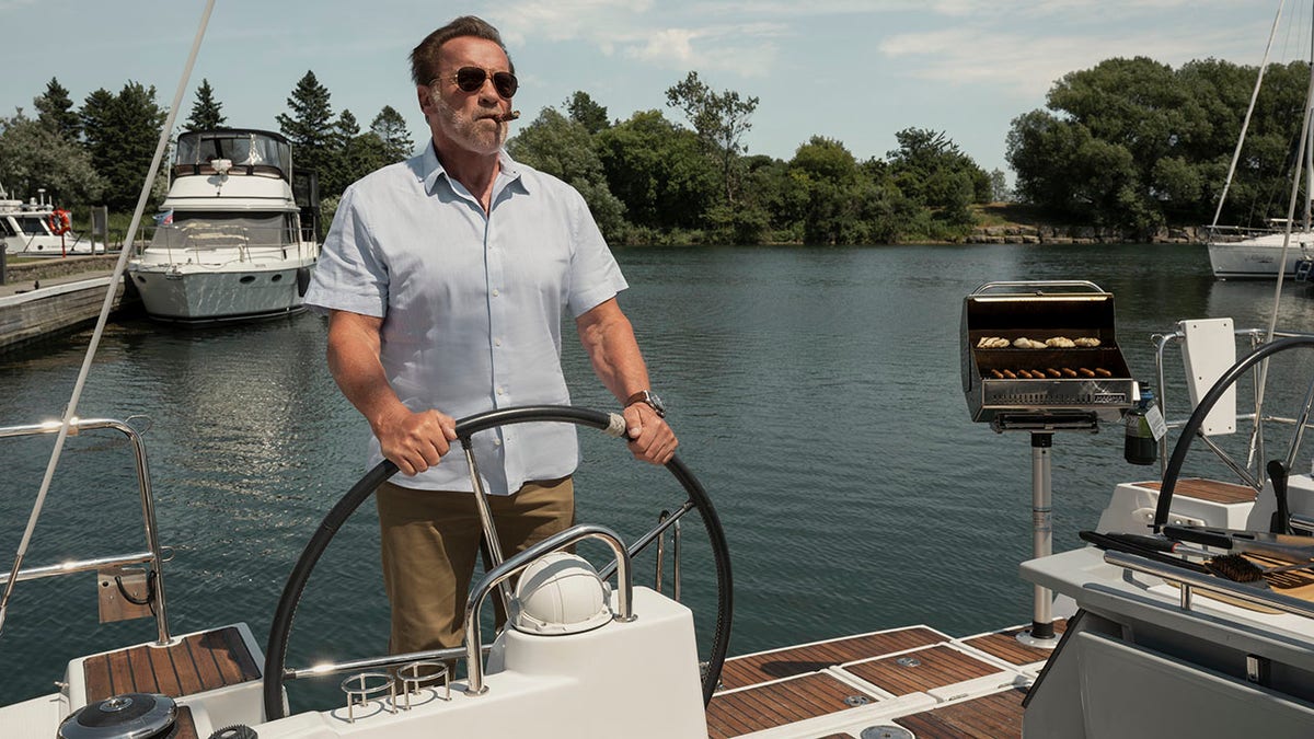 Arnold Schwarzenegger in a light blue button-down short sleeve and khaki pants steers a large boat and smokes a cigar in "FUBAR"