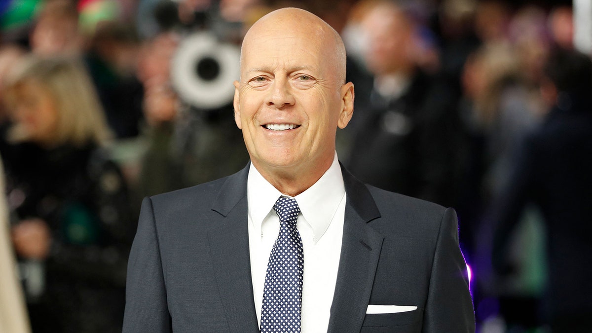 Bruce Willis smiles on the red carpet wearing a black suit, a white button down and a blue tie