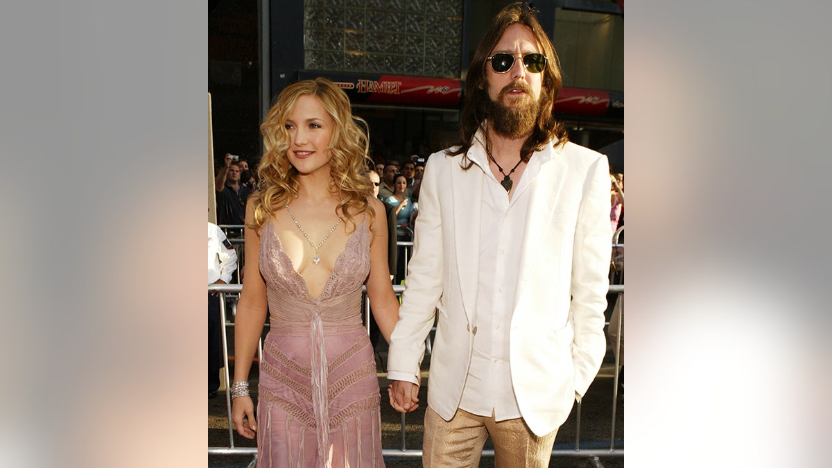 Kate Hudson in a plunging light pink lacy dress holds hands with Chris Robinson in a white jacket and tan flair pants on the red carpet