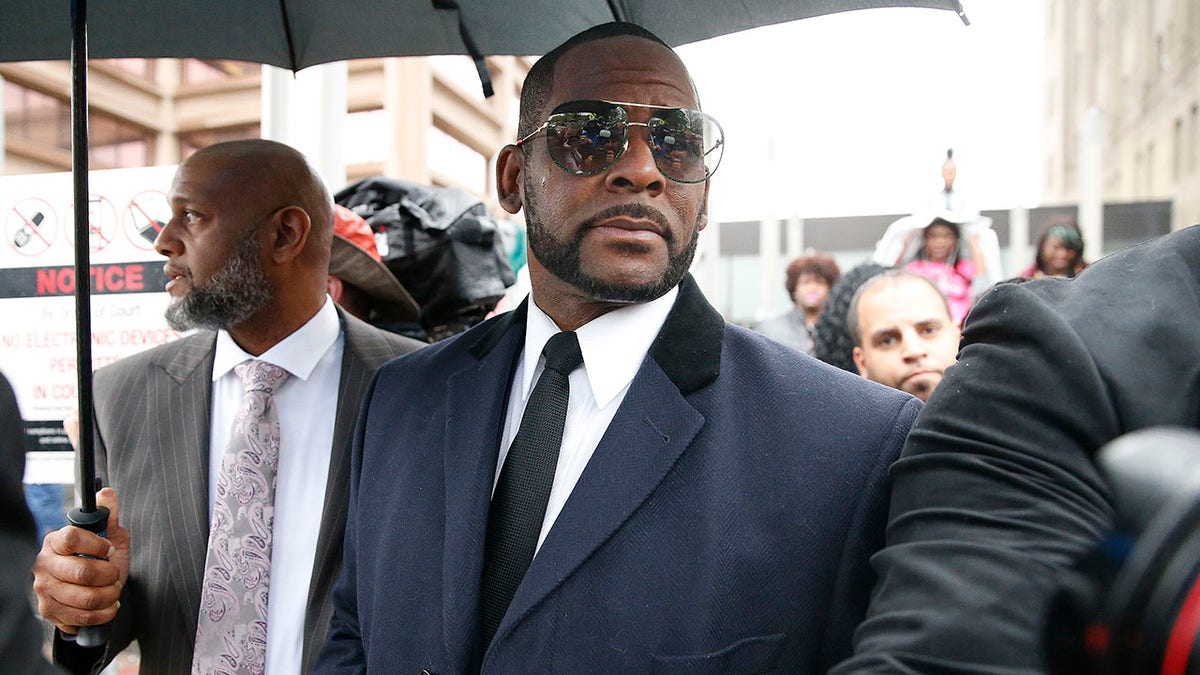 R. Kelly in a blue jacket with a black collar and black tie and dark shades leaves the courthouse