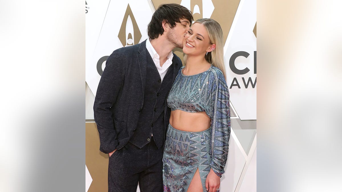 Morgan Evans in a black suit and vest and white shirt kisses Kelsea Ballerini in a sequined blue two-piece set on the CMA red carpet in 2019