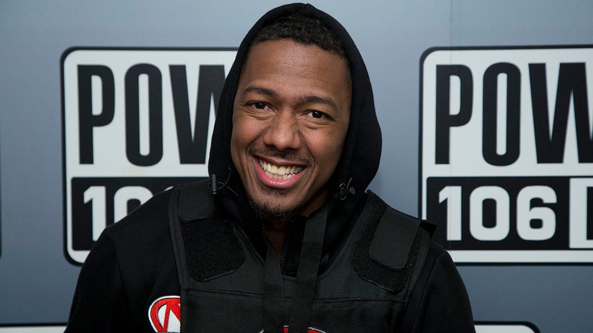 Nick Cannon smiles in a black hooded sweatshirt and vest