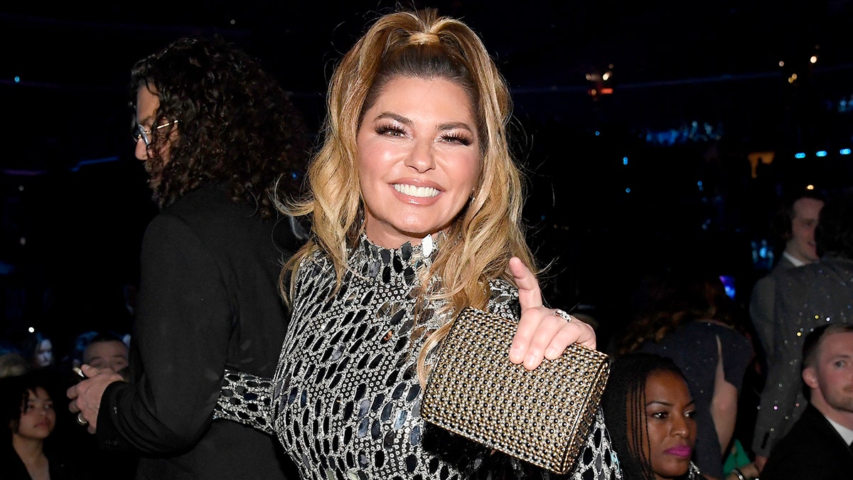 Shania Twain in a black and white jeweled dress with a half-up half-down ponytail and sparkly bag in her hand smiles at the camera