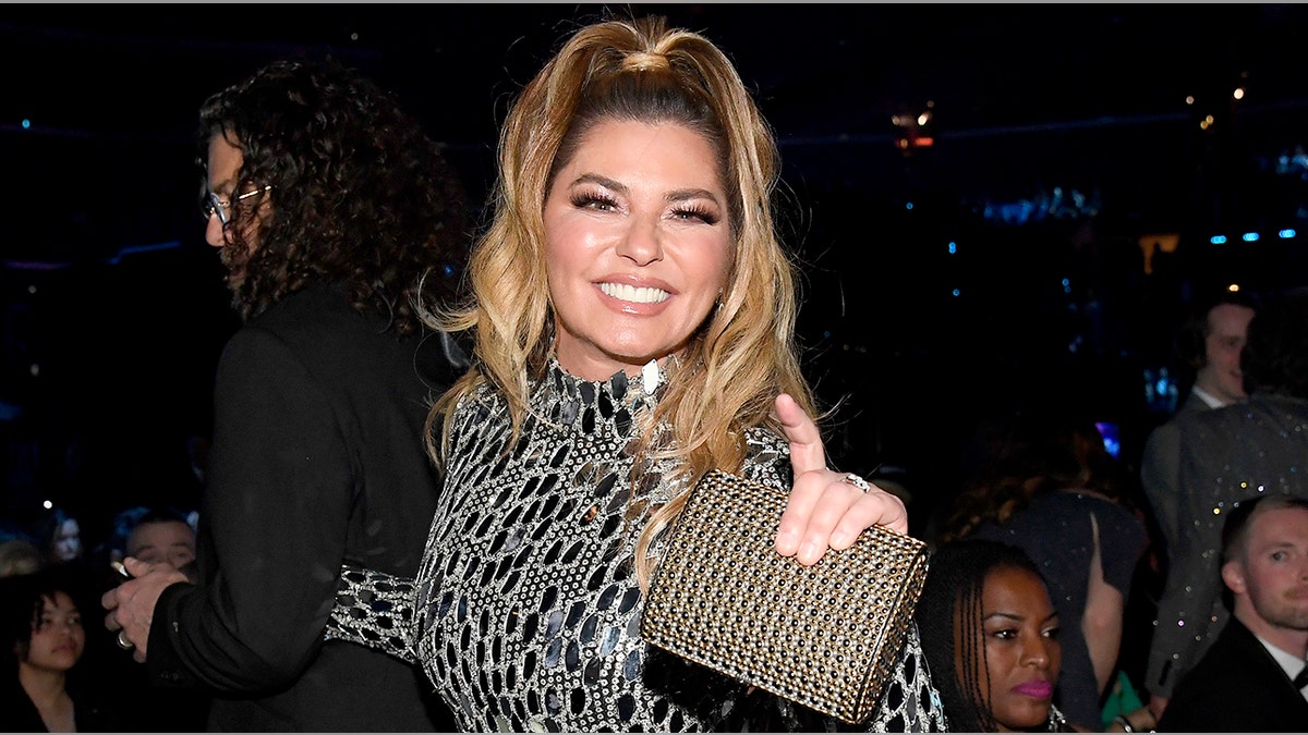 Shania Twain in a black and white jeweled dress with a half-up half-down ponytail and sparkly bag in her hand smiles at the camera