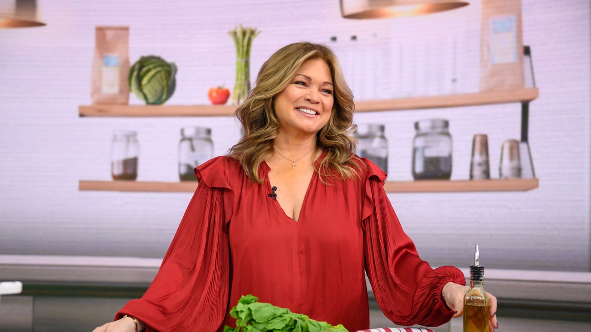Valerie Bertinelli wears a red shirt and stands behind a counter with food.