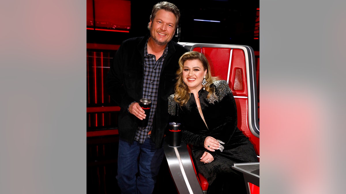 Blake Shelton in a plaid shirt and black suit smiles behind Kelly Clarkson in a black outfit sitting in her coaching chair on "The Voice"