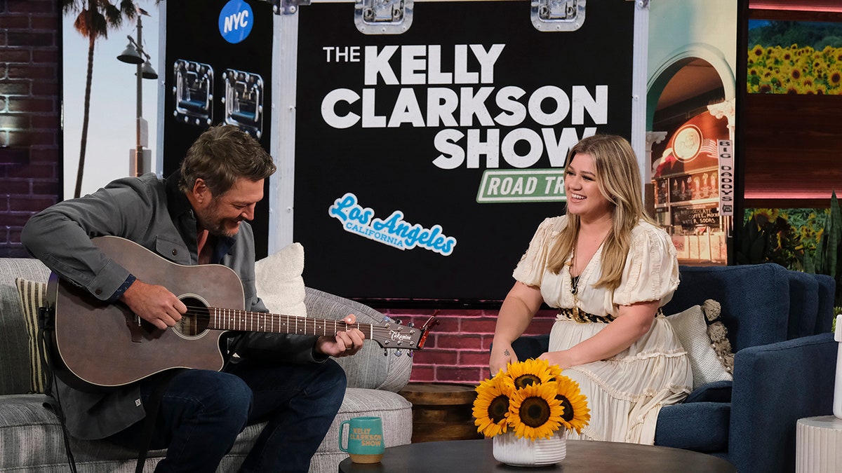 Kelly Clarkson in a white long dress looks on during her talk show while guest Blake Shelton strums on the guitar on her couch