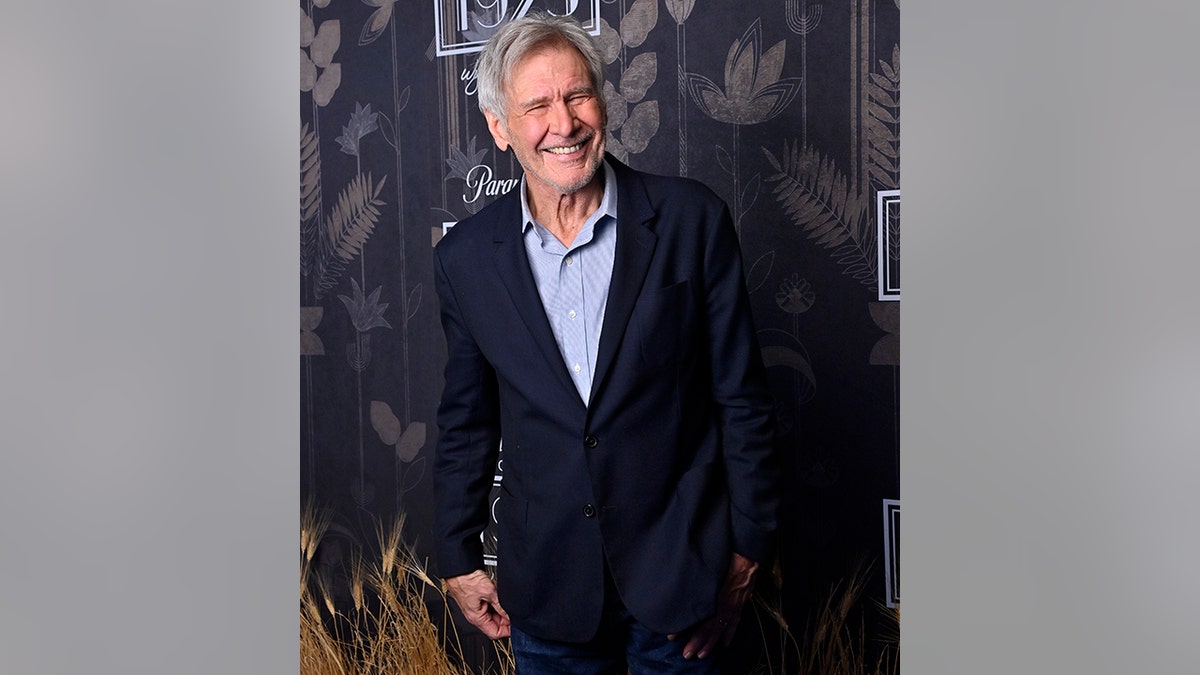 Harrison Ford smiles in a light blue button down and navy suit at the premiere of "1923"