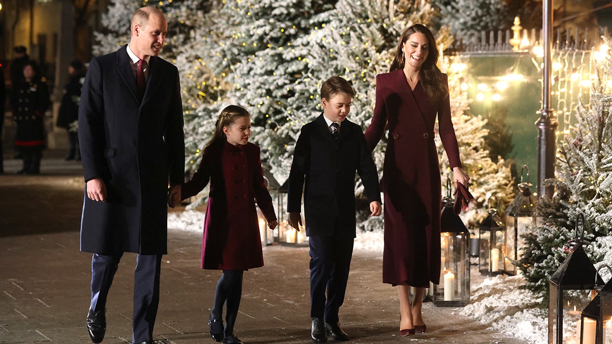 Prince William in a black coat and maroon tie holds hands with Princess Charlotte in a maroon jacket next to Prince George in a black jacket, being led by mother Kate Middleton in a maroon long jacket for a Christmas Carol service on Dec. 15