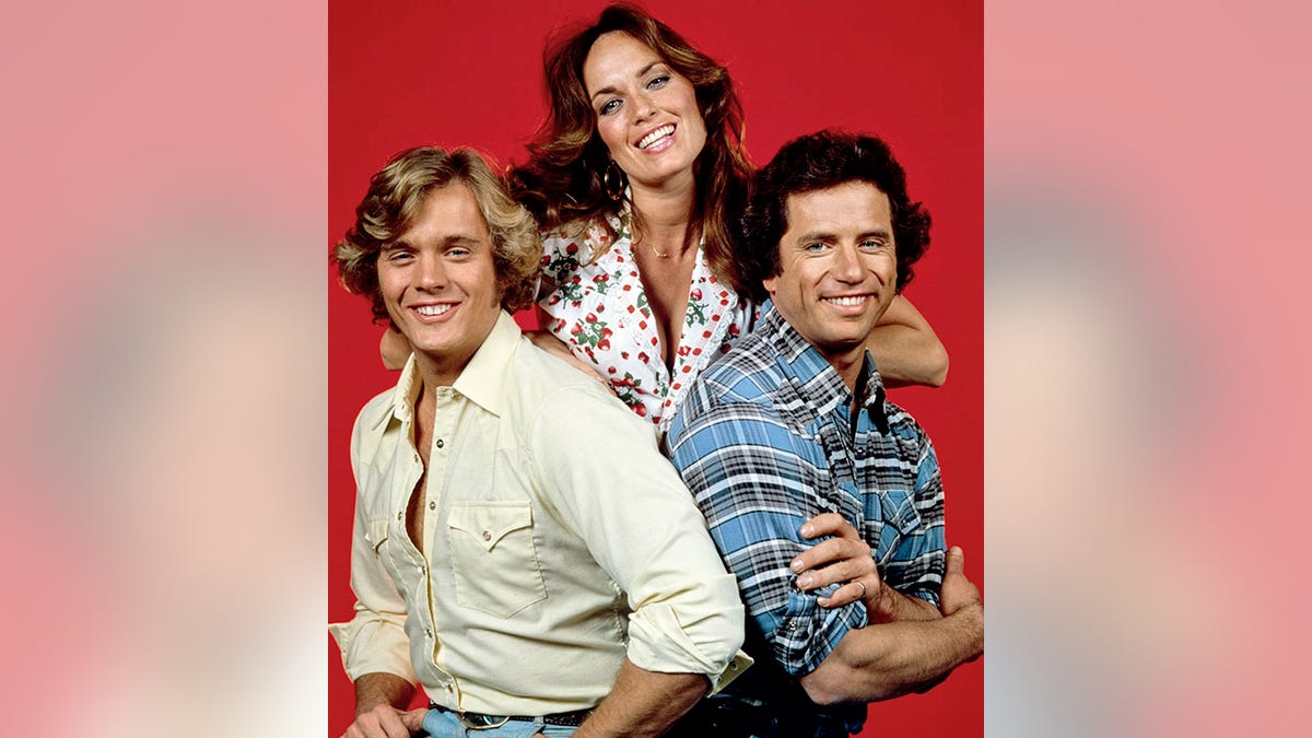 John Schneider as Bo Duke in a white button down shirt smiles next to Catherine Bach as Daisy Duke in a white printed top, next to Tom Wopat as Luke Duke in a blue plaid shirt in a photo for "The Dukes of Hazzard"