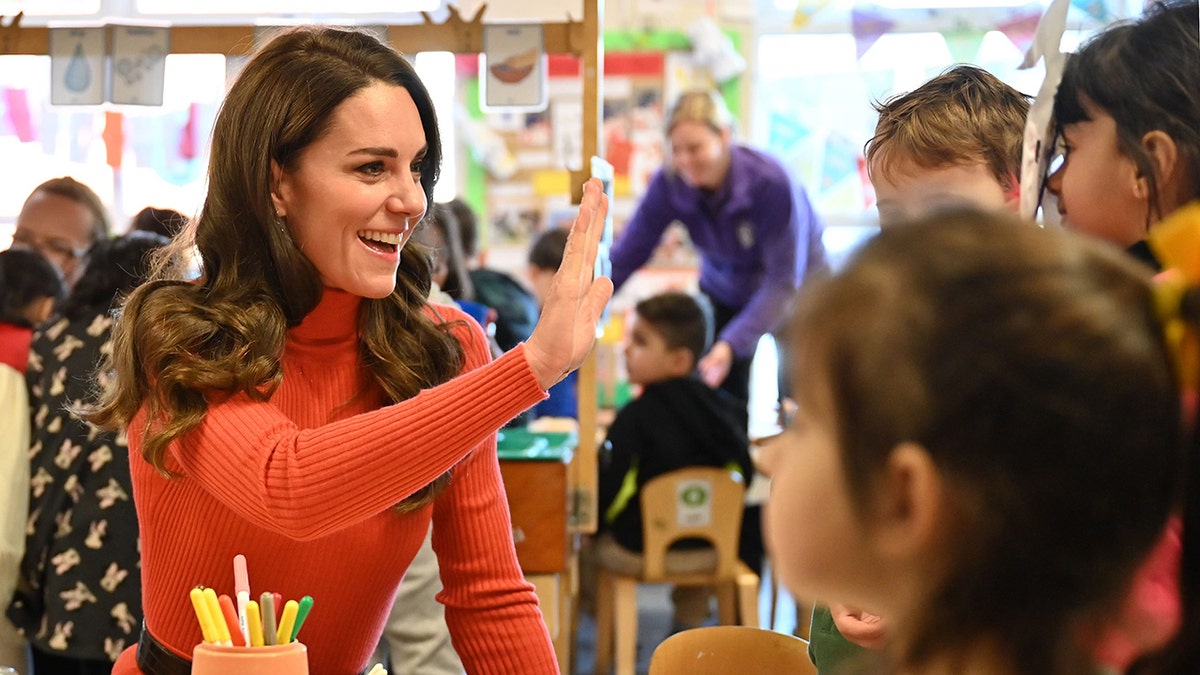 Kate Middleton in an orange turtleneck reaches out for a high-five in a nursery with children