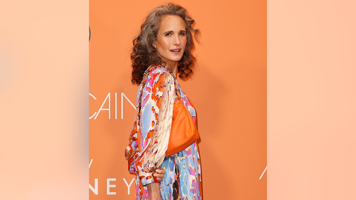 Andie MacDowell on the red carpet is photographed mid-turn with a colorful printed dress and bright orange bag at the Marc Cain Fashion show in Berlin