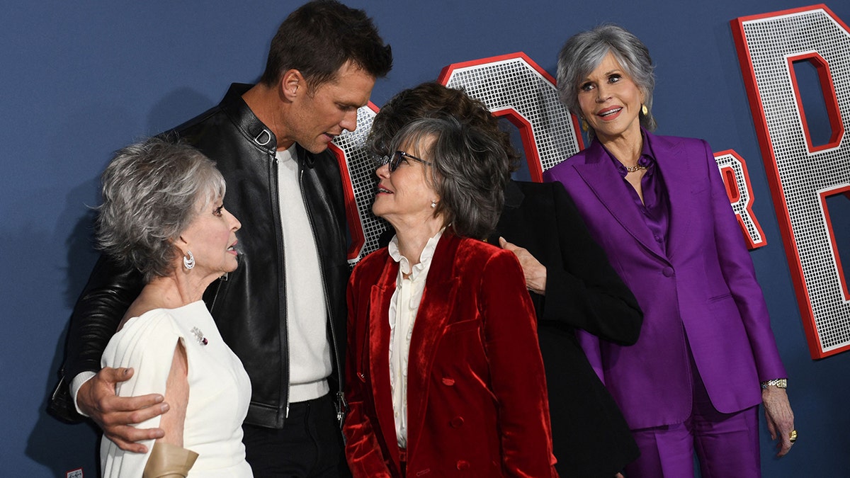 Rita Moreno looks at Tom Brady who hugs both her and Sally Field, with Jane Fonda looking on in a purple suit, and Lily Tomlin trying to pop in behind Sally Field