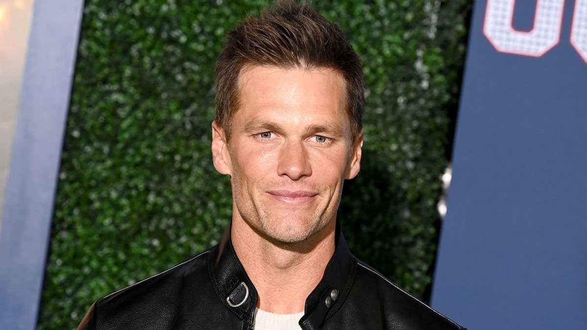 Tom Brady in a white shirt and leather jacket soft smiles and stares right at the camera on the "80 For Brady" red carpet