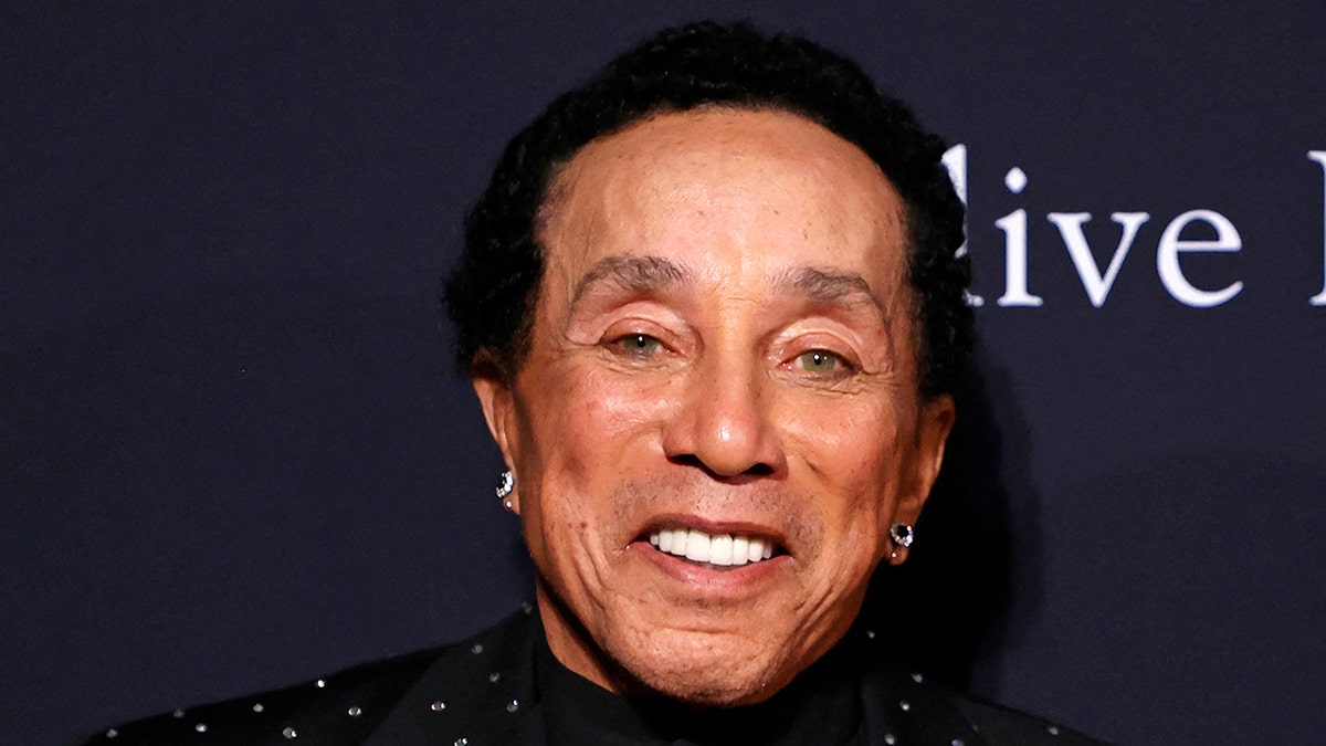 Smokey Robinson smiles on the red carpet in a black sparkly suit and earrings