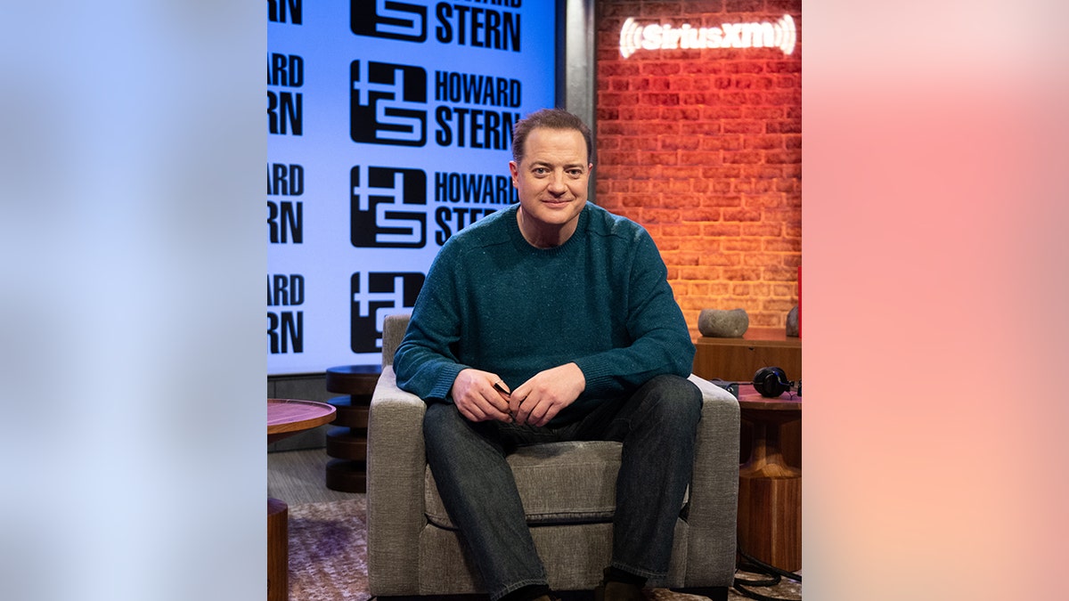 Brendan Fraser sits in a grey chair with a dark teal shirt on in the studio for The Howard Stern show
