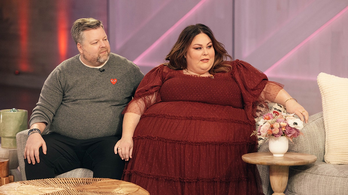 Chrissy Metz in a long maroon ruffled dress rests her hand on her boyfriend Bradley Collin's knee wearing a grey sweater while on "The Kelly Clarkson Show"