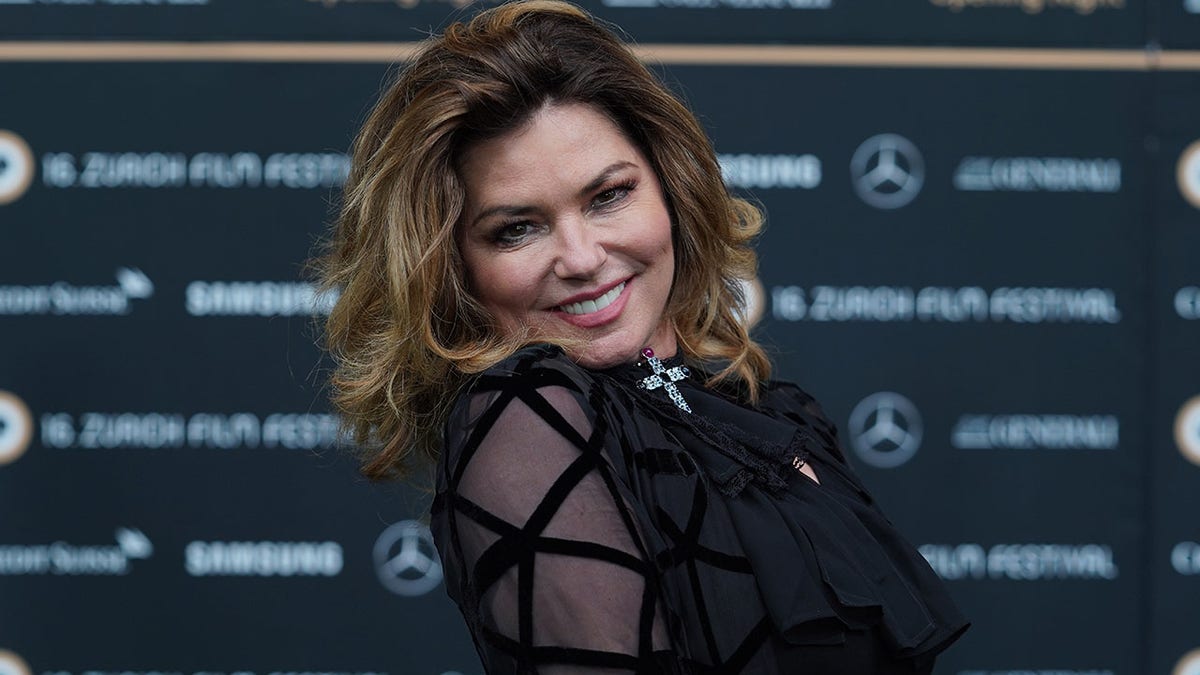 Shania Twain in a black outfit with sheer criss-cross sleeves smiling on the carpet