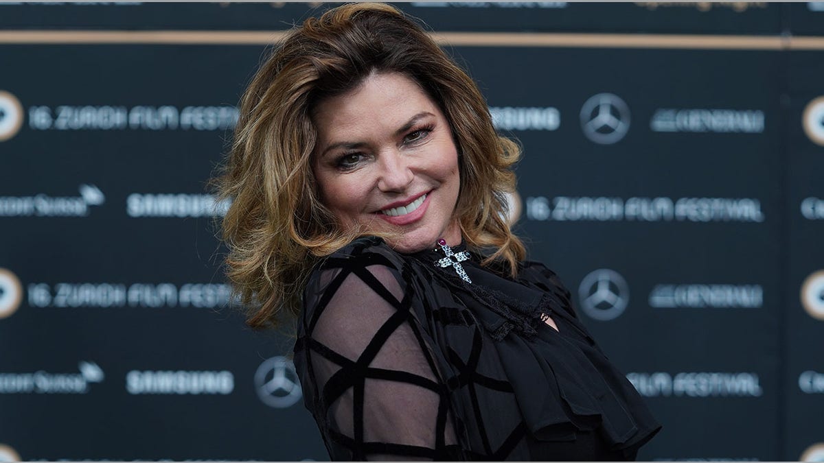 Shania Twain in a black outfit with sheer criss-cross sleeves smiling on the carpet