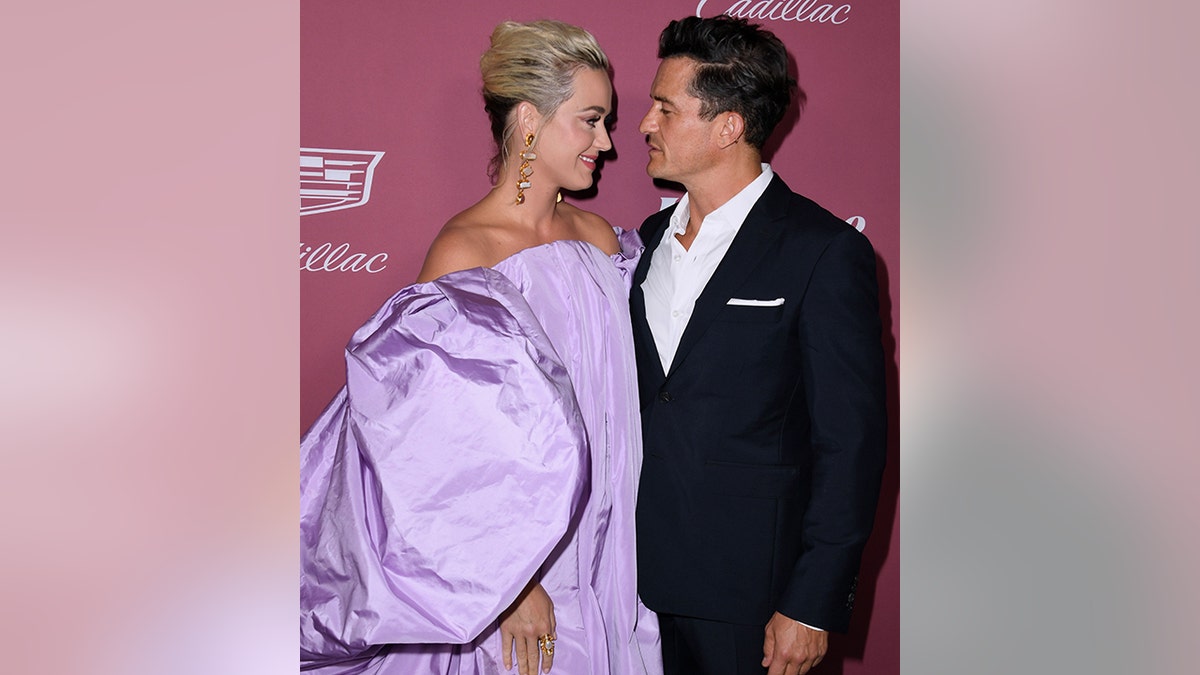 Katy Perry in a large fluffy lavender dress stares and smiles at Orlando Bloom in a white button down and black jacket on the red carpet