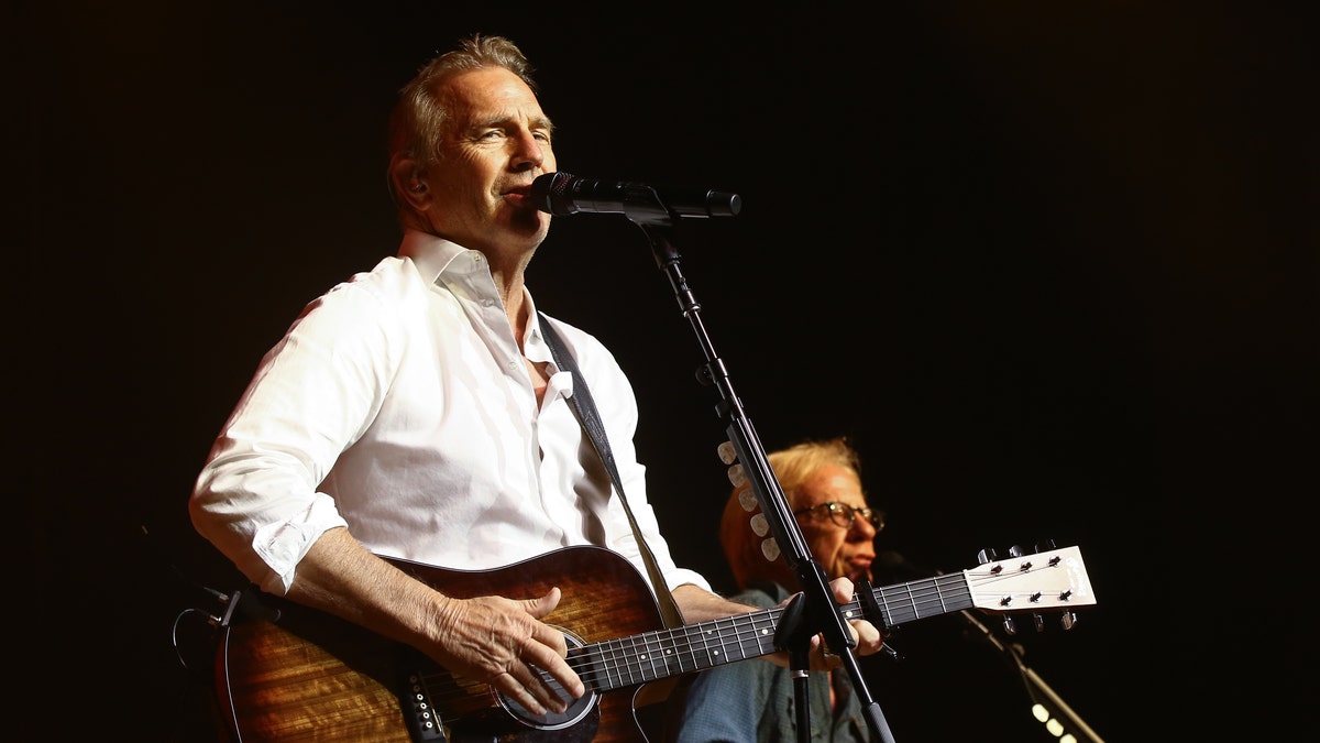 Kevin Costner holds a guitar and wears a white shirt while performing on stage
