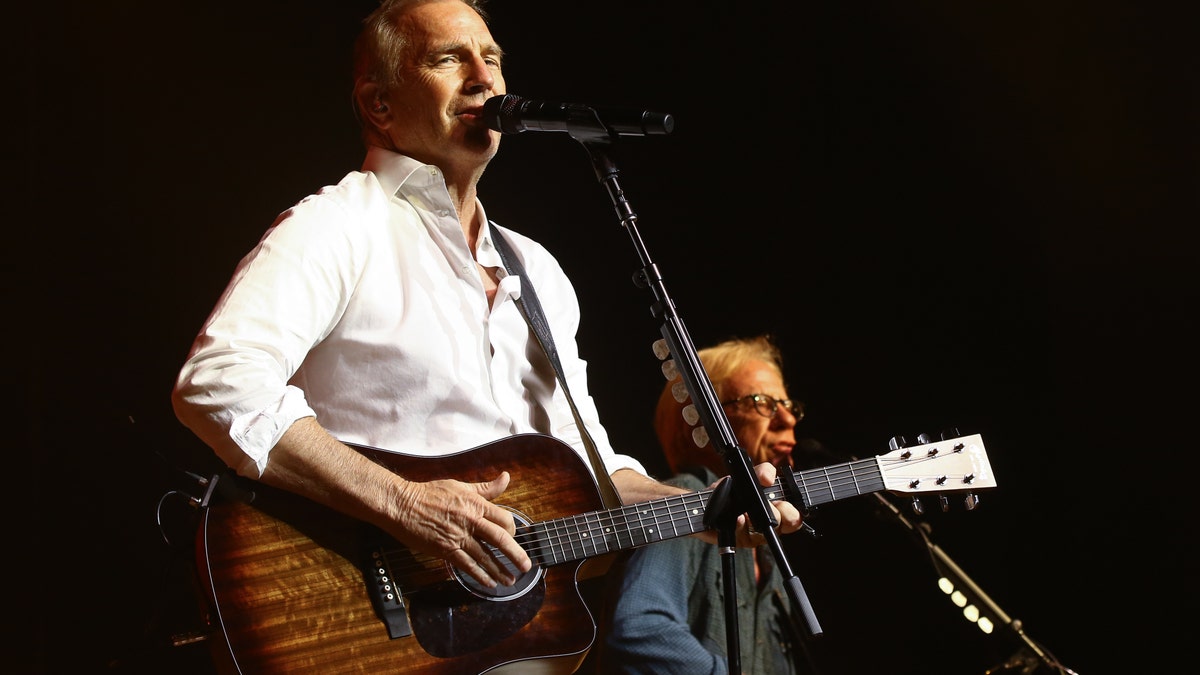 Kevin Costner holds a guitar and wears a white shirt while performing on stage