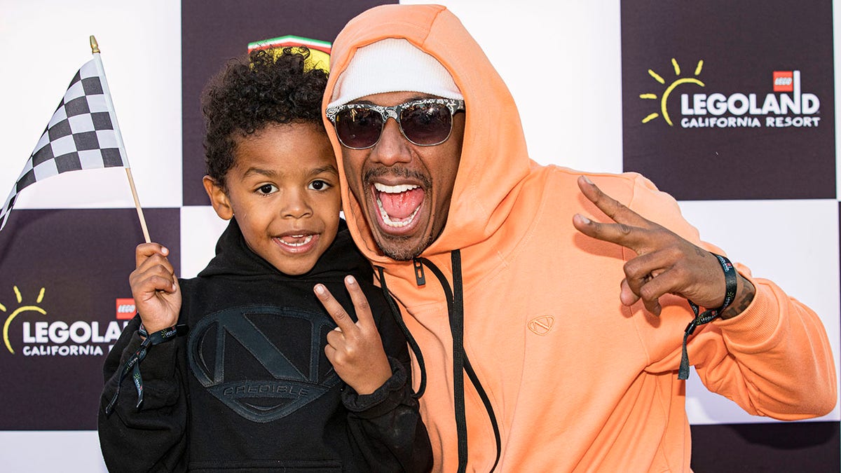 Nick Cannon in an orange sweatshirt, white band around his head and dark sunglasses poses with his son Golden Cannon doing a peace sign while wearing black on the red carpet