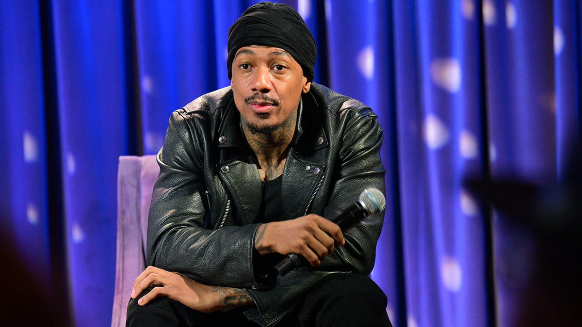 Nick Cannon with a black turban leans over in a black leather jacket sitting on a purple chair holding a microphone