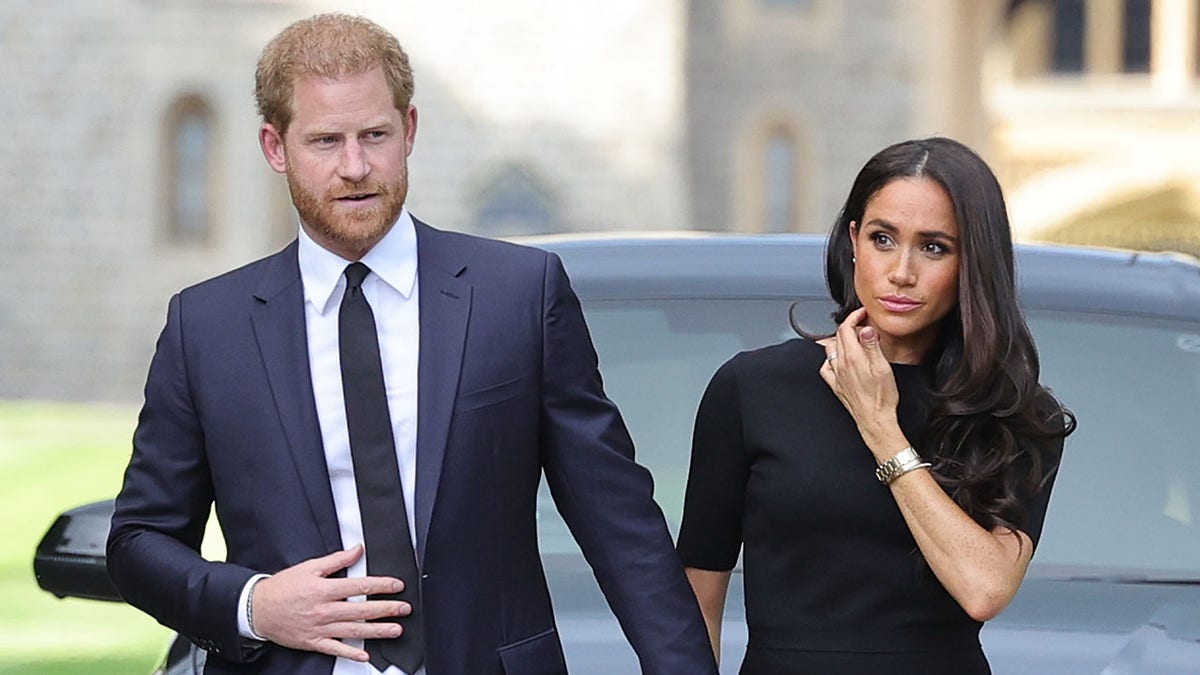 O Príncipe Harry de terno azul-marinho e gravata segura a mão de Meghan Markles, usando um vestido escuro após o falecimento da Rainha