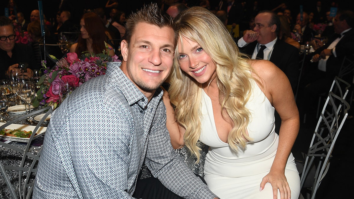 Rob Gronkowski in a blue patterned button down smiles next to Camille Kostek in a white dress